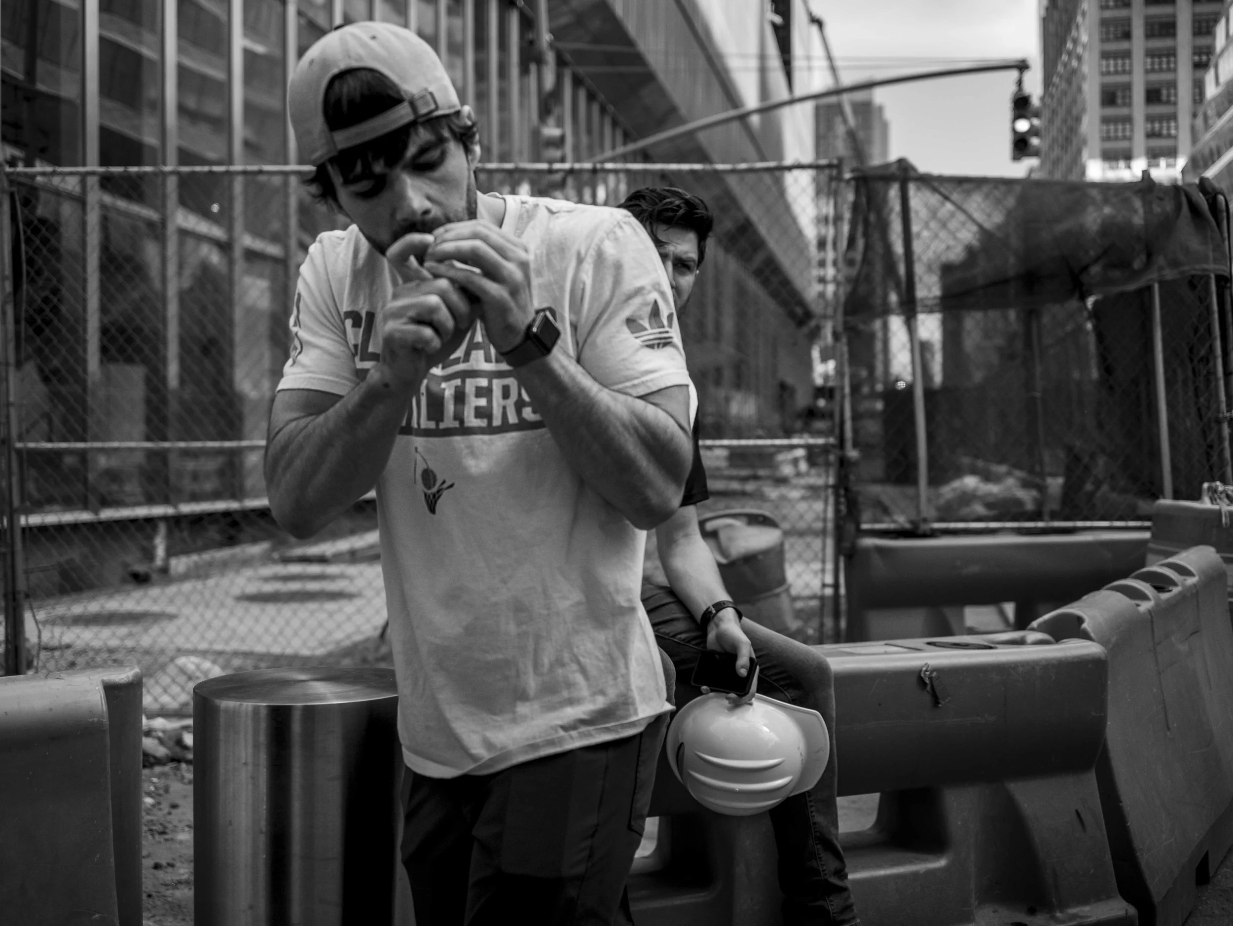 Two young men sitting outdoors in an urban area, one is wearing a cap backwards and looking down, holding a cellphone, the other is behind him, partially visible, with a serious expression. There are metal barricades, a fence, and tall buildings in t