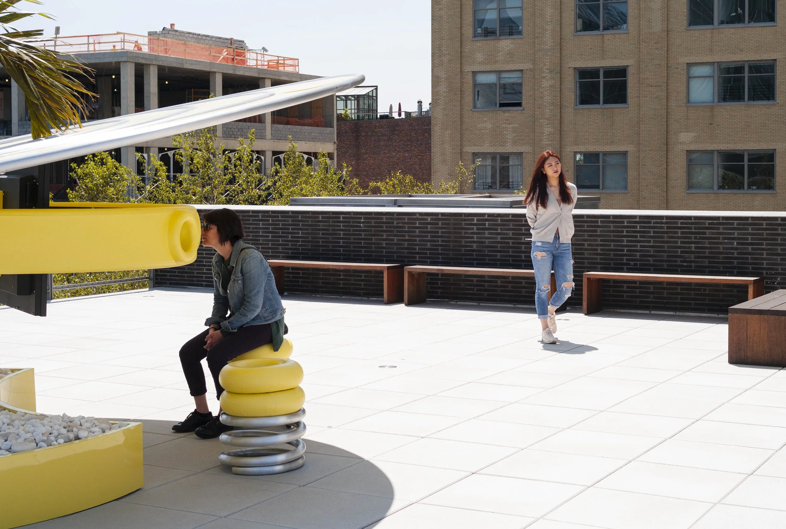 Two women on a rooftop terrace with modern buildings in the background. One woman with dark hair and sunglasses sits on a yellow seating sculpture made of stacked circular shapes; the other woman with long brown hair, wearing a white jacket with ripp