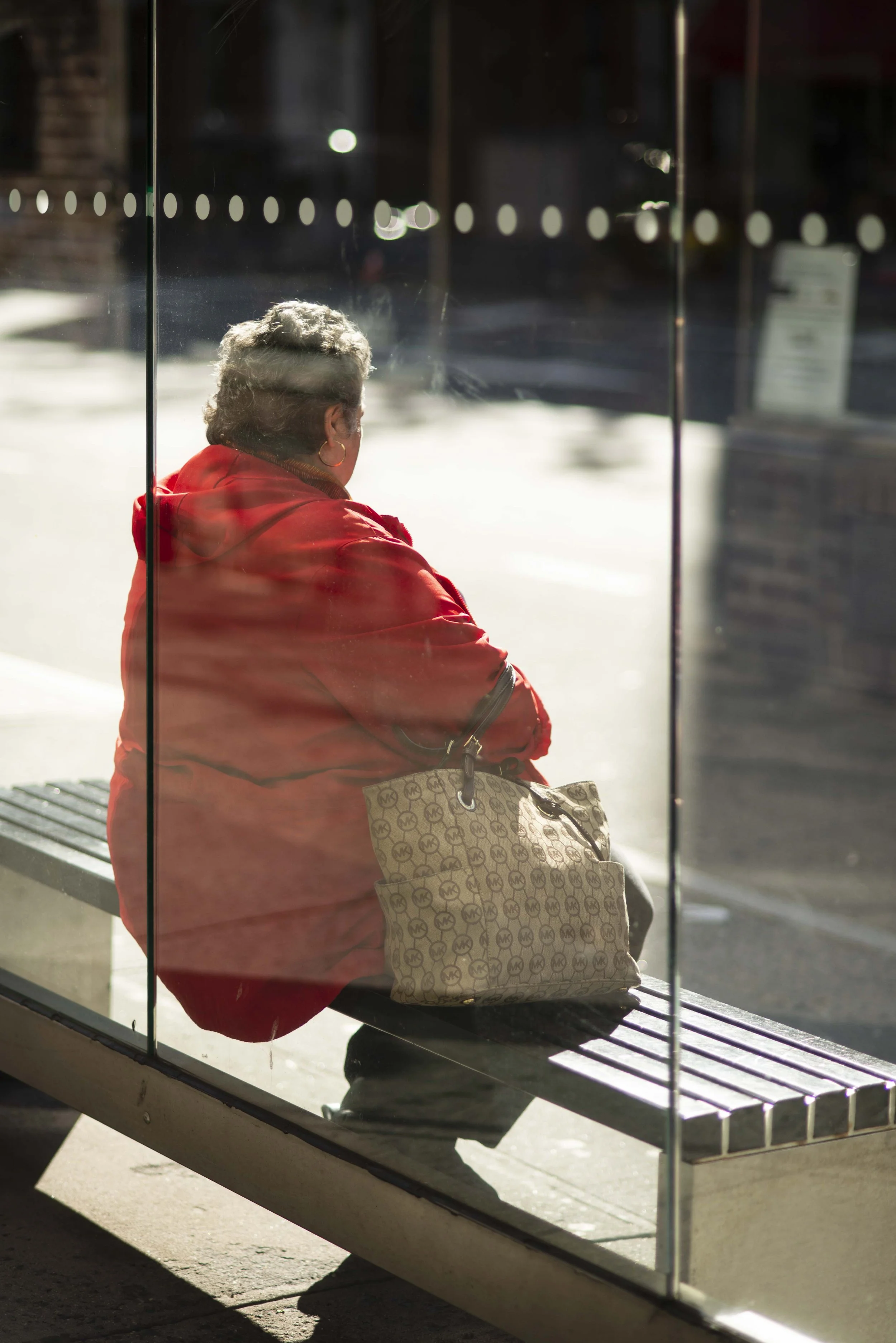 An older woman with short gray hair and gold hoop earrings sitting at a bus stop, wearing a red jacket and holding a Michael Kors handbag, seen through the glass of the bus shelter.