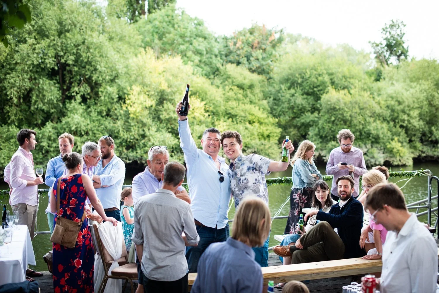 Group of people celebrating outdoors near a river with trees in the background, holding drinks, smiling, and socializing at a gathering.