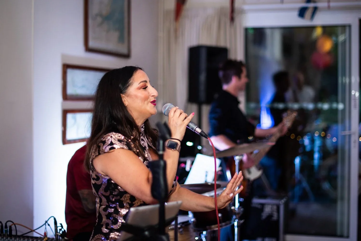 Woman singing into a microphone with musicians playing instruments in the background at an indoor event.