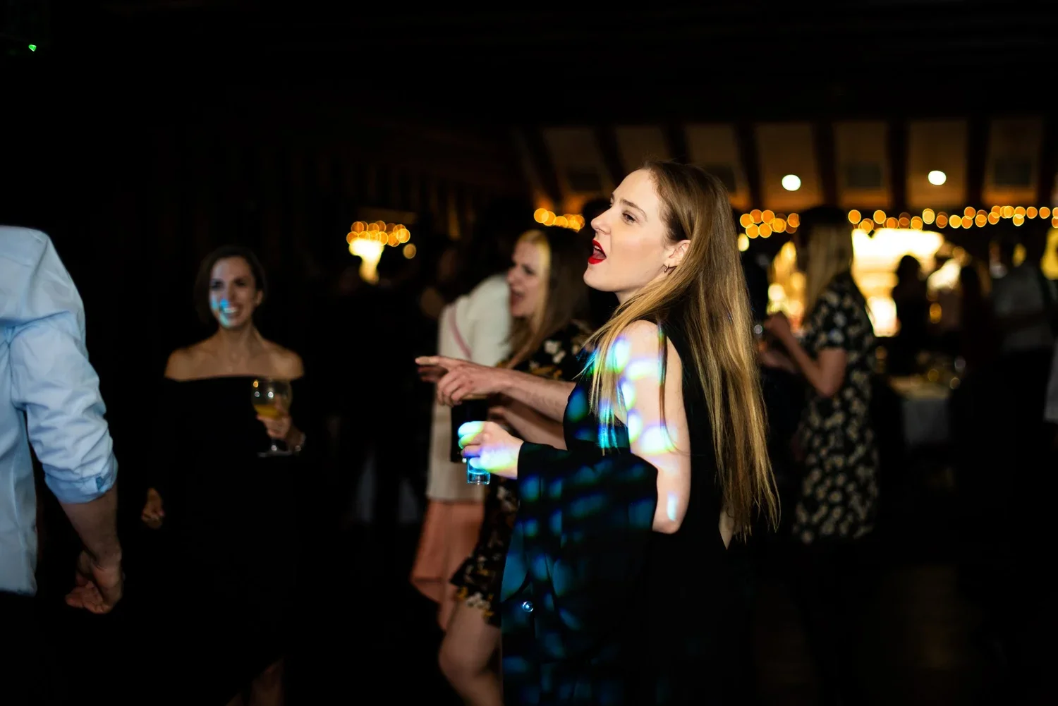 People dancing and socializing at an indoor party with string lights, with women holding drinks and enjoying the music.