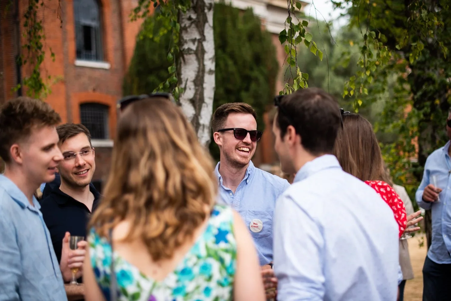 A group of people socializing outdoors during daytime, smiling and engaging in conversation with trees and brick buildings in the background.