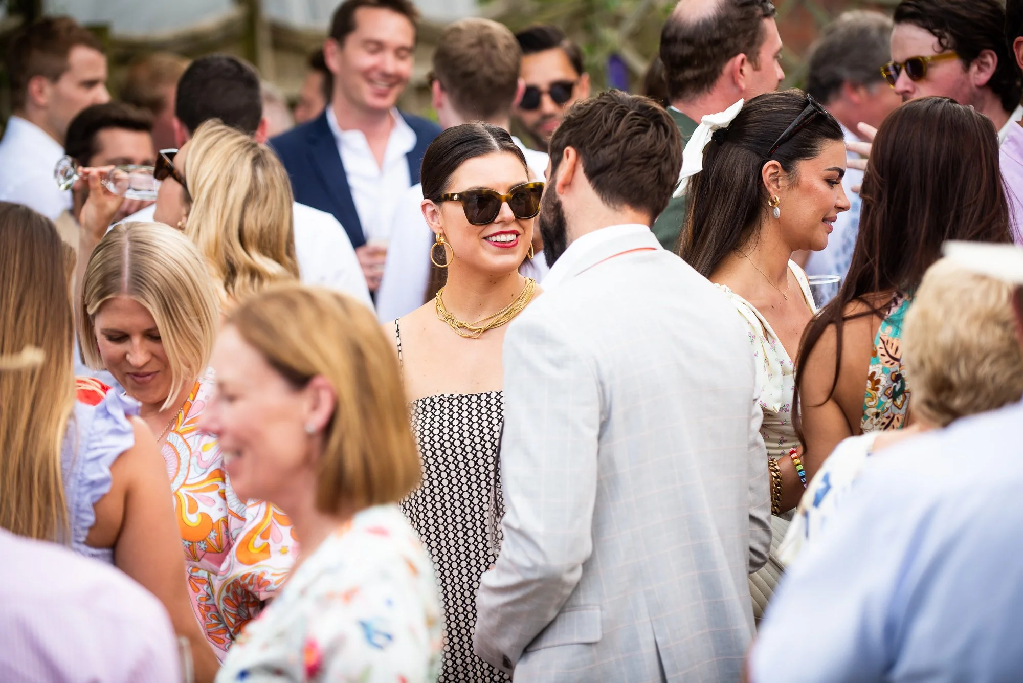 A group of people at an outdoor social gathering, dressed in summer attire, engaging in conversation and smiling.