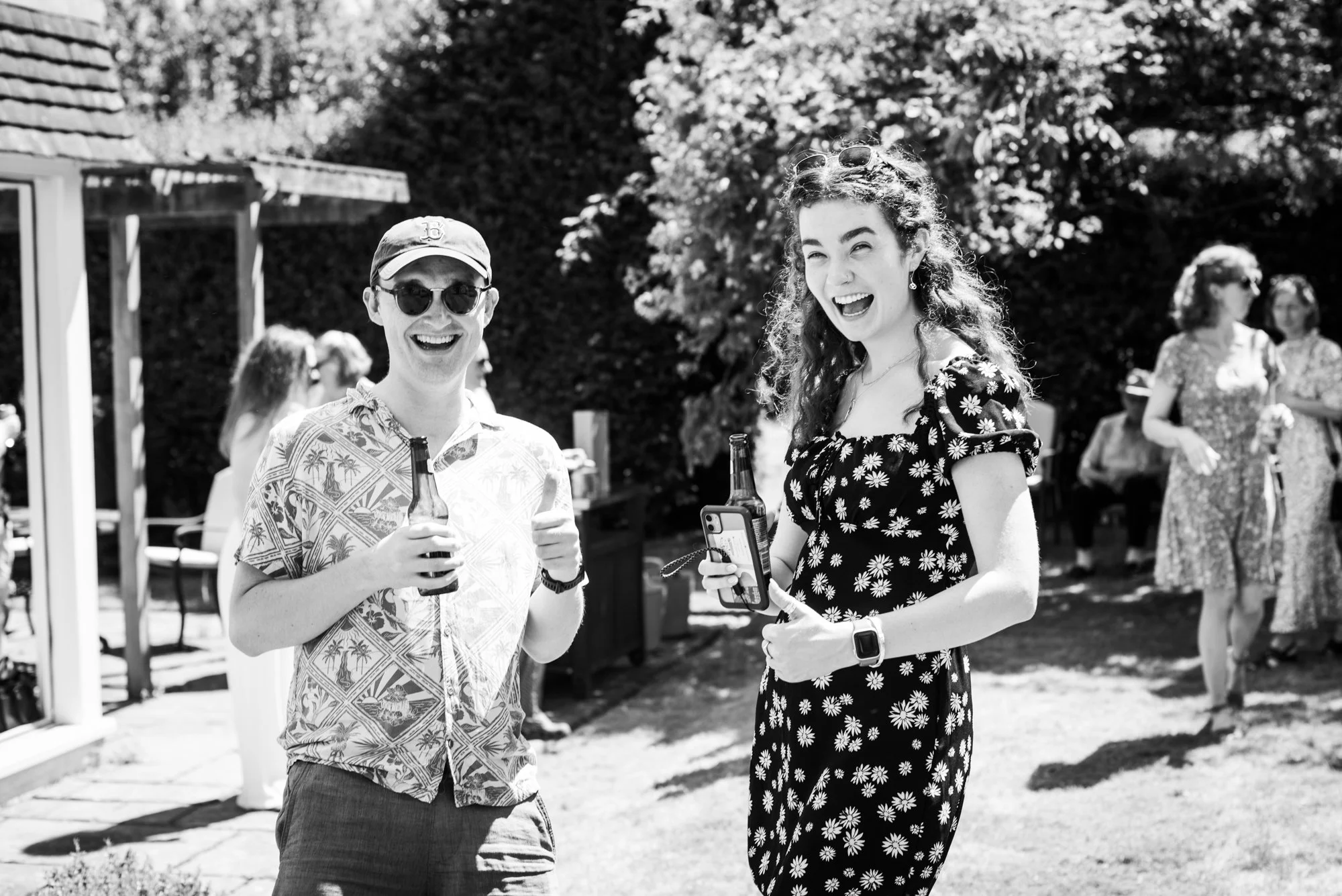 Two young people at an outdoor gathering, smiling and holding drinks, with other guests in the background.