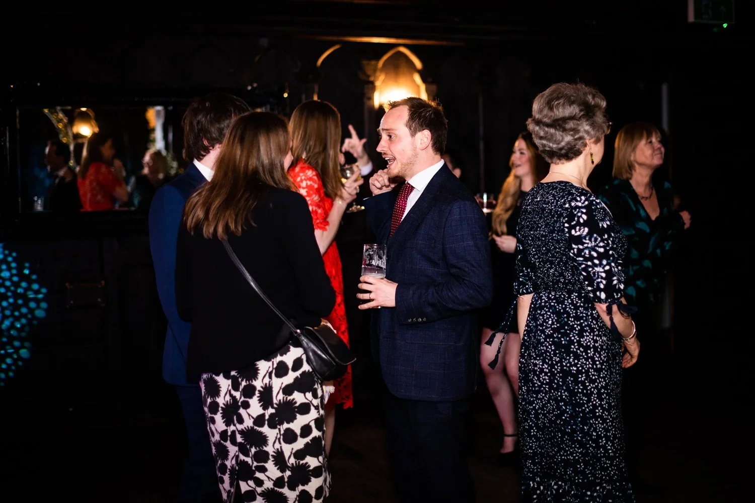 People socializing at a party or event in a dimly lit room, some holding drinks, engaged in conversation.