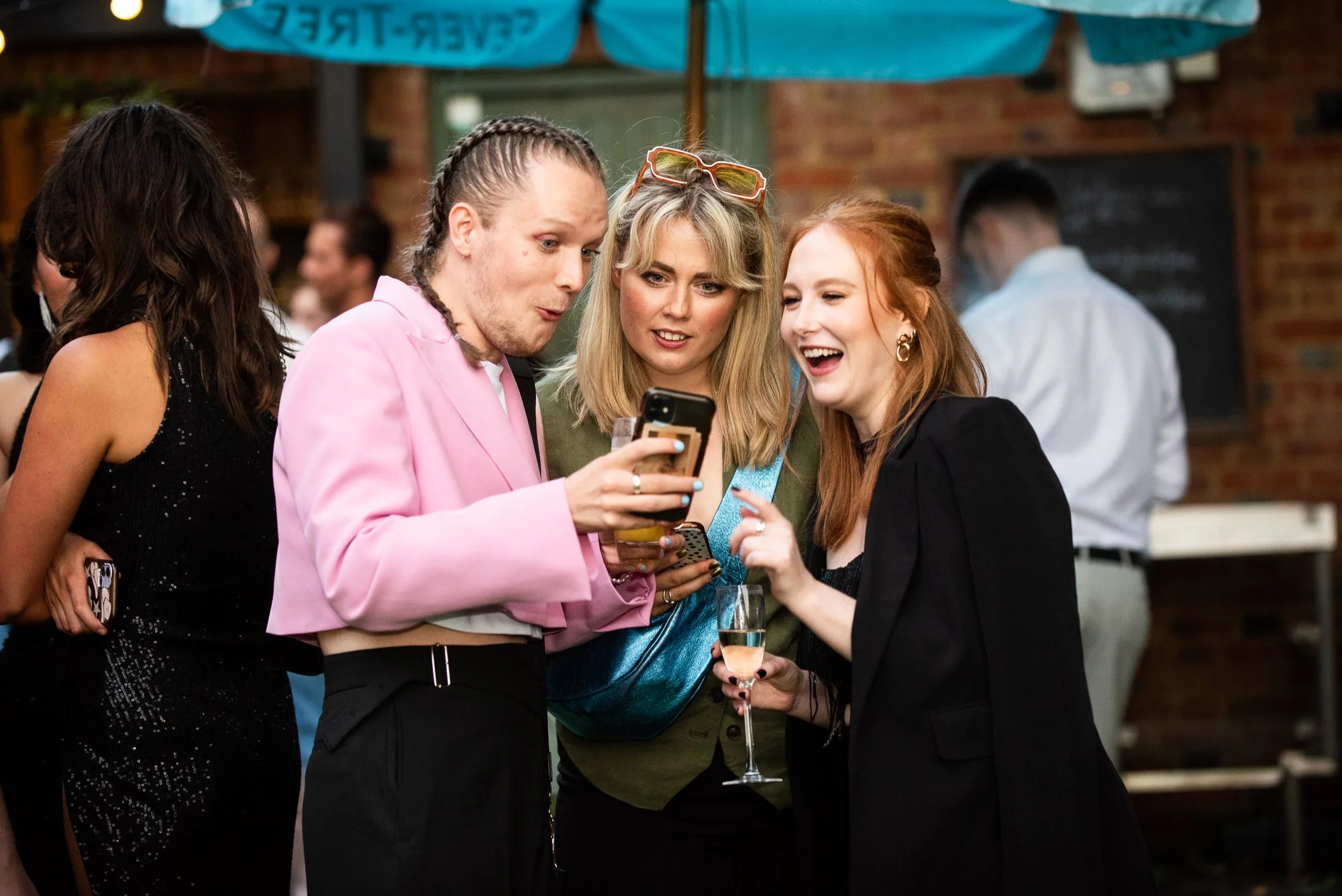 Group of four young women at a social gathering, looking at a smartphone and smiling, with drinks in hand, indoors with a brick wall background.