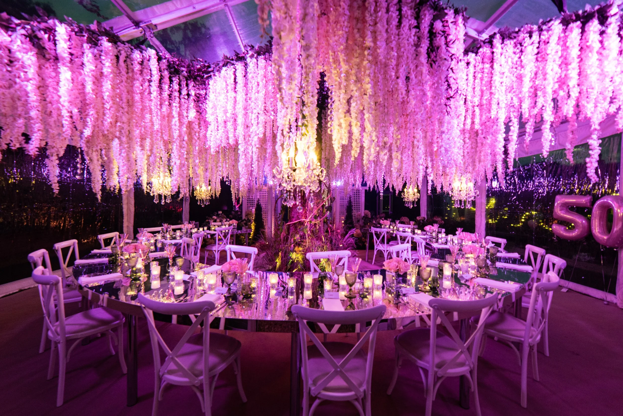 Elegant banquet hall decorated with pink and purple floral arrangements, hanging flowers from the ceiling, and a central tree wrapped in flowers, with round tables, white chairs, candles, and balloons in the background.