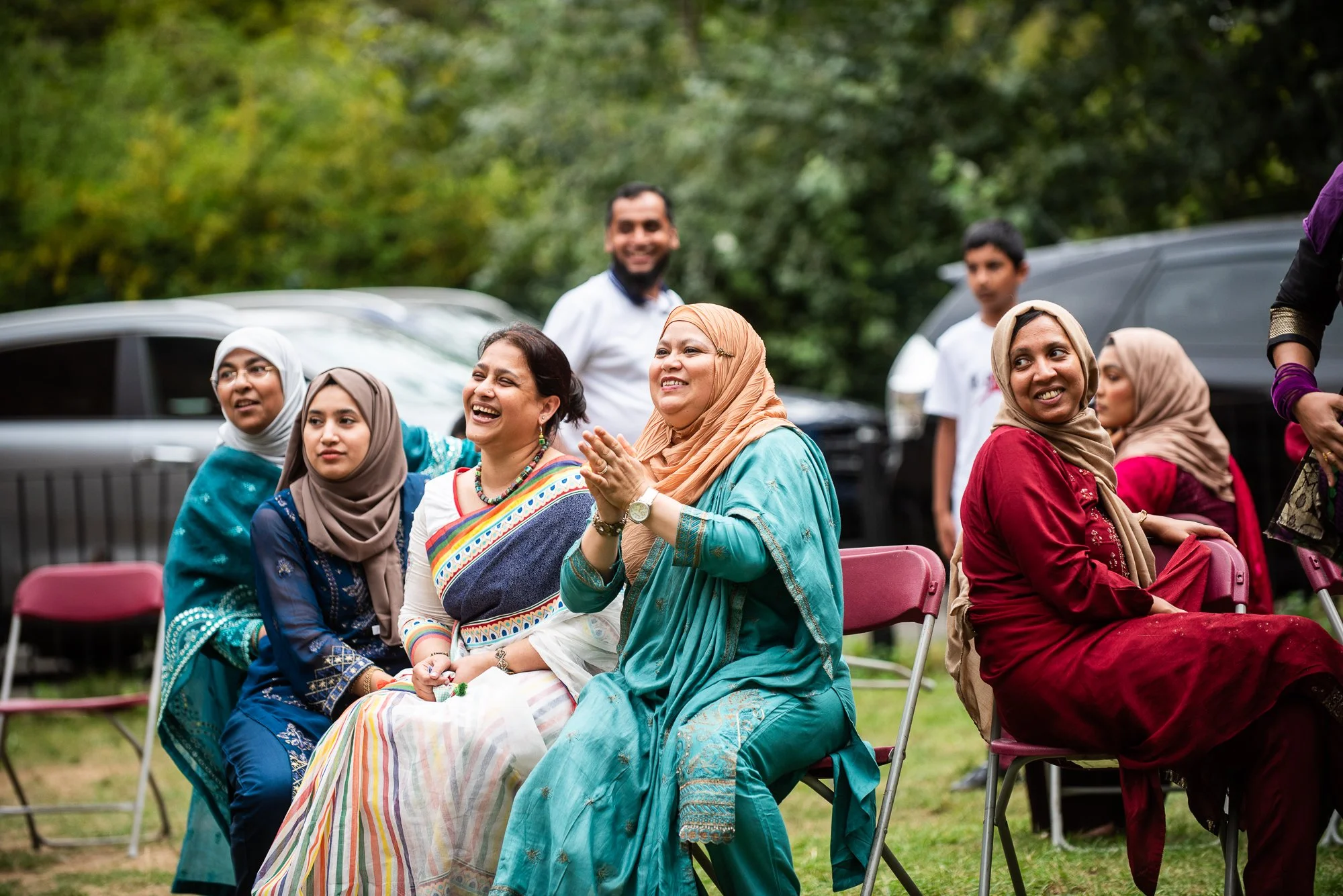 Group of diverse women and men sitting and standing outdoors, smiling and enjoying an event amid greenery.