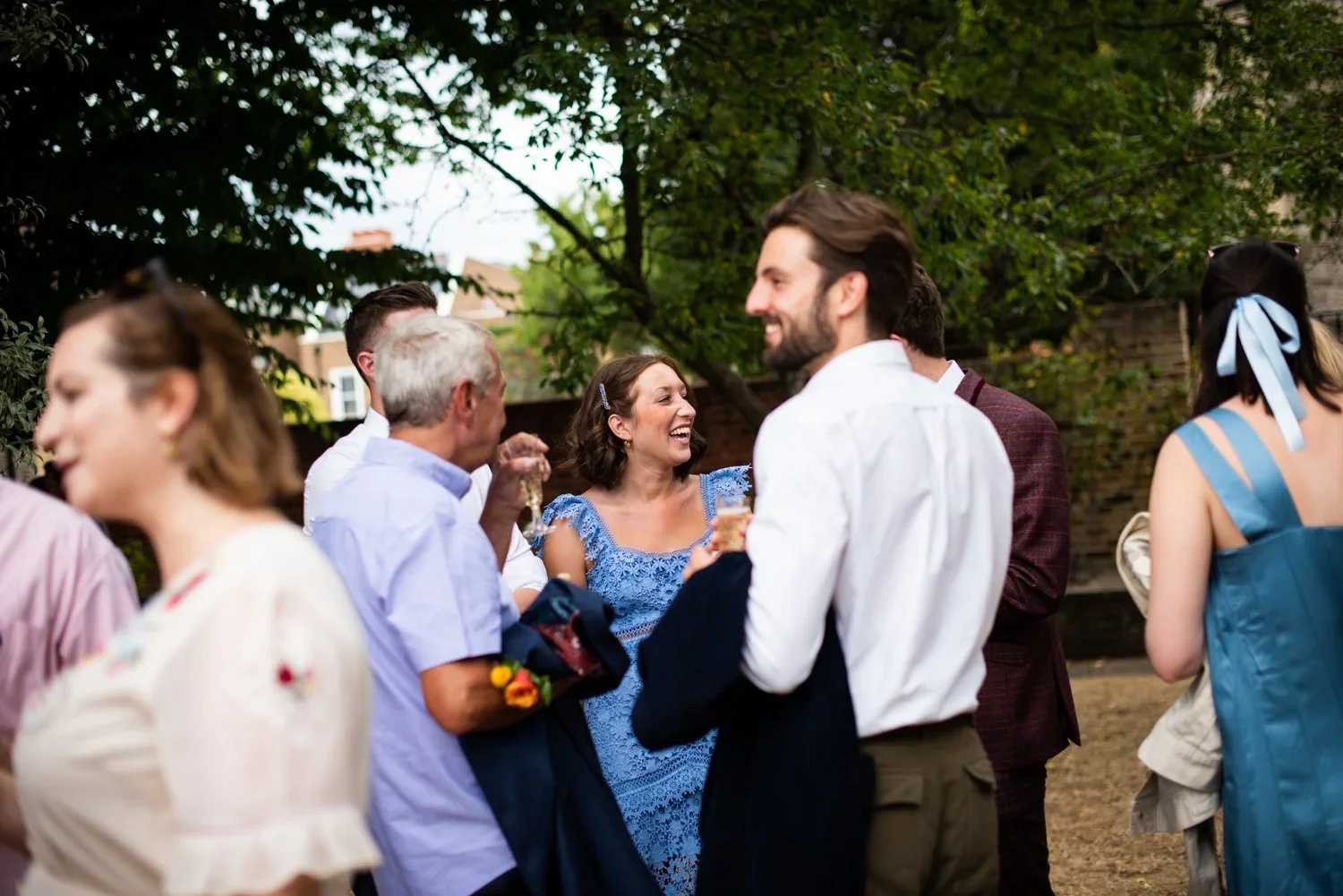 A group of people at an outdoor celebration or party, talking and laughing among trees and greenery.