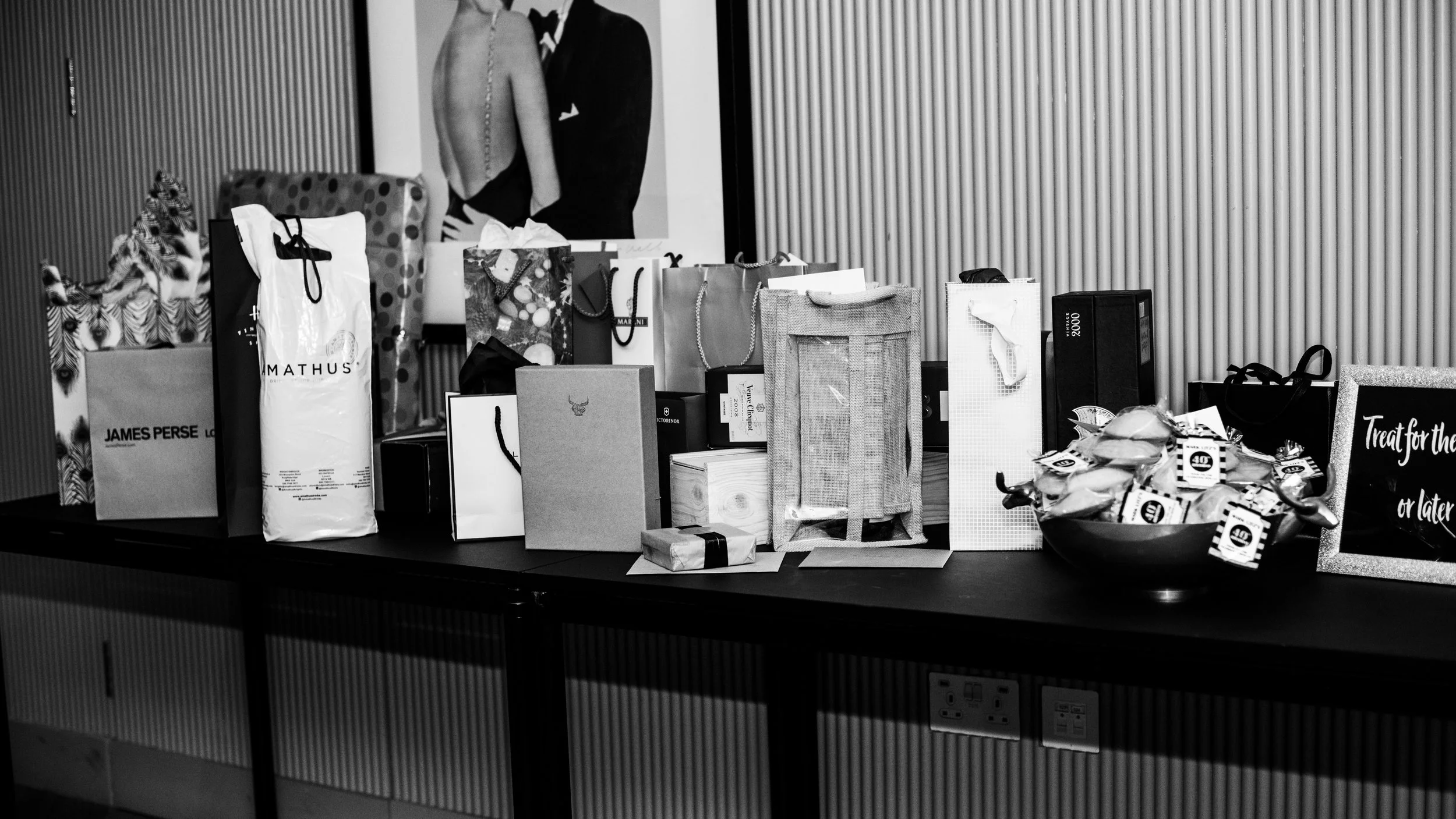 Table with various gift bags, boxes, and wrapped presents, with a framed black and white artwork of people in formal attire in the background.