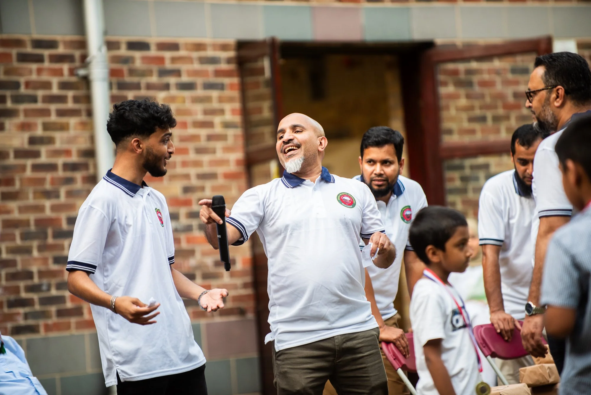 A man with a bald head and beard holding a microphone, smiling and talking to a young man with curly hair, in an outdoor setting with a brick wall background, surrounded by several people including a young boy and adults engaged in a conversation.