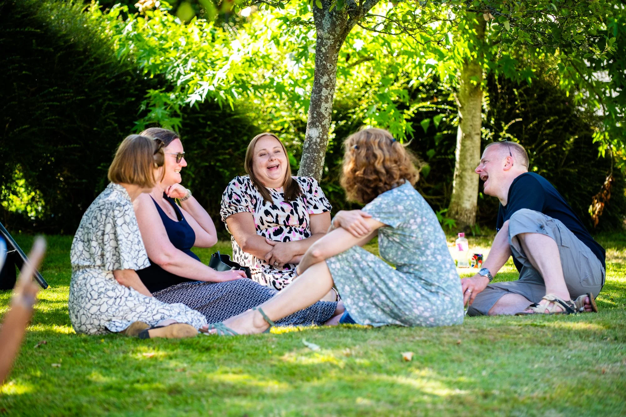 Five people sitting on the grass in a park, smiling and laughing, enjoying a sunny day.
