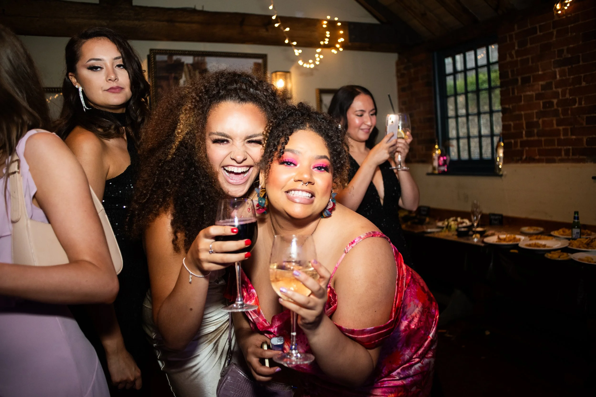 Group of five women celebrating at a party indoors, smiling and holding wine glasses, with two women in the background taking photos. The room has warm lighting, exposed brick walls, and a long table with food.