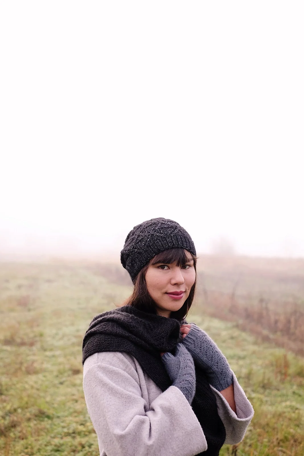 Dark haired woman wearing hand knit textured hat, shawl and mitts in a foggy field.