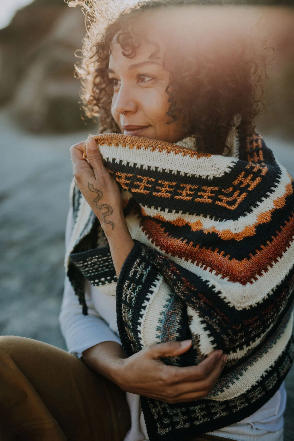 Curly haired woman wearing multi coloured mosaic knit triangular shawl on boulder in front of ocean.