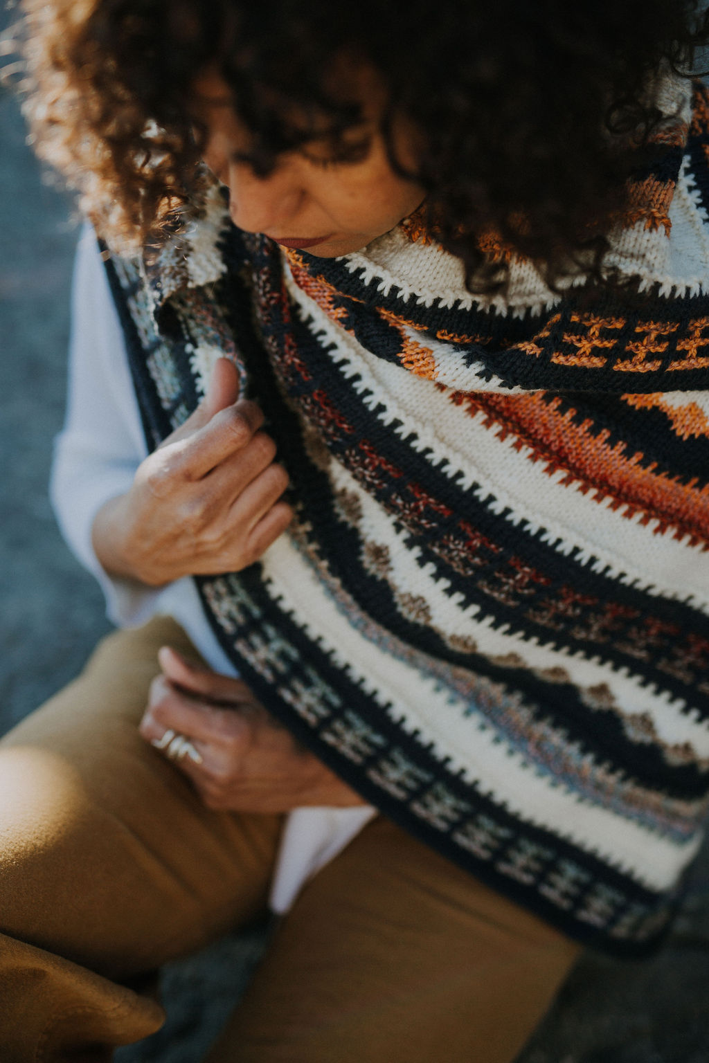 Curly haired woman wearing multi coloured mosaic knit triangular shawl on boulder in front of ocean in various poses.