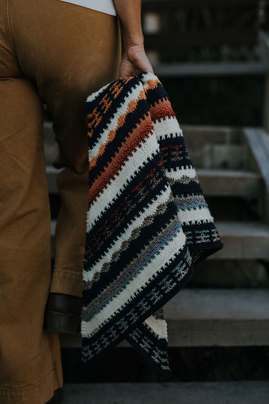 Curly haired woman wearing multi coloured mosaic knit triangular shawl on boulder in front of ocean in various poses.