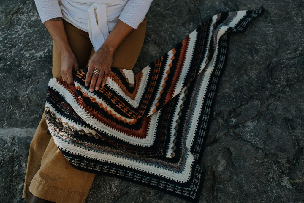 Curly haired woman wearing multi coloured mosaic knit triangular shawl on her lap on boulder in front of ocean.