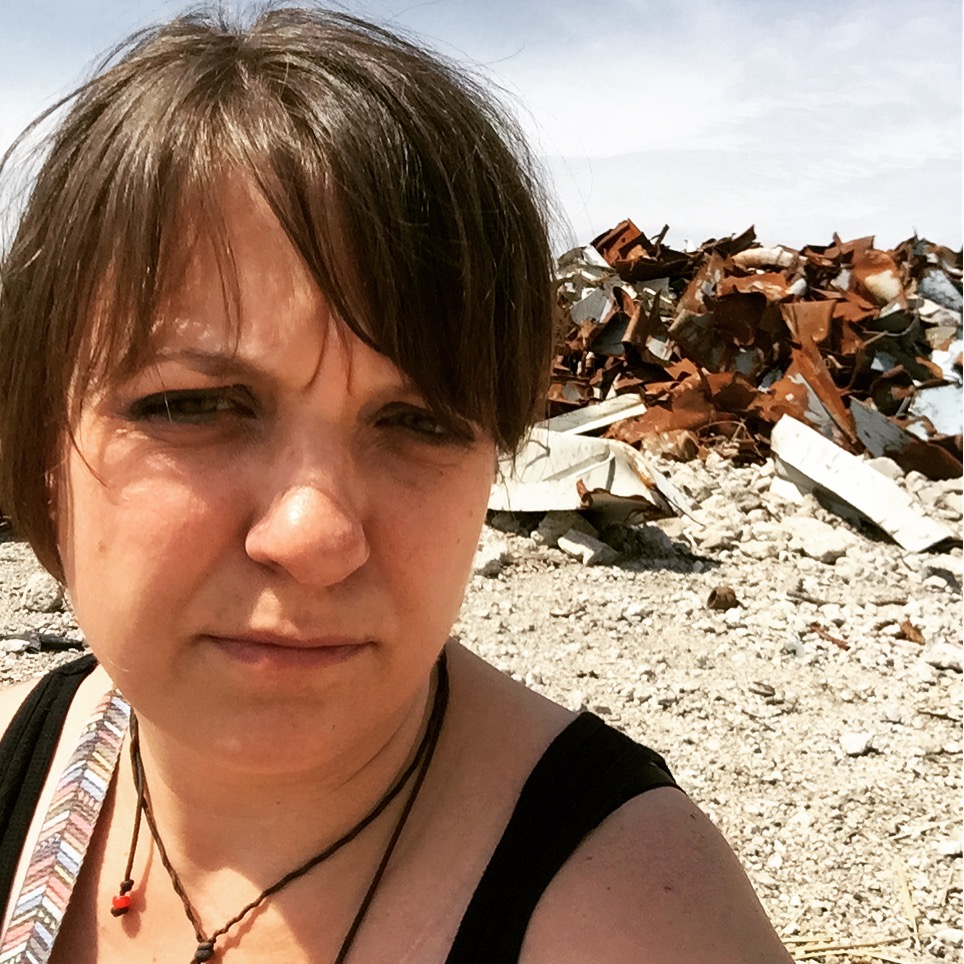 A woman with short brown hair and a black tank top taking a selfie outdoors against a background of scattered debris and scrap metal on a rocky terrain under a partly cloudy sky.