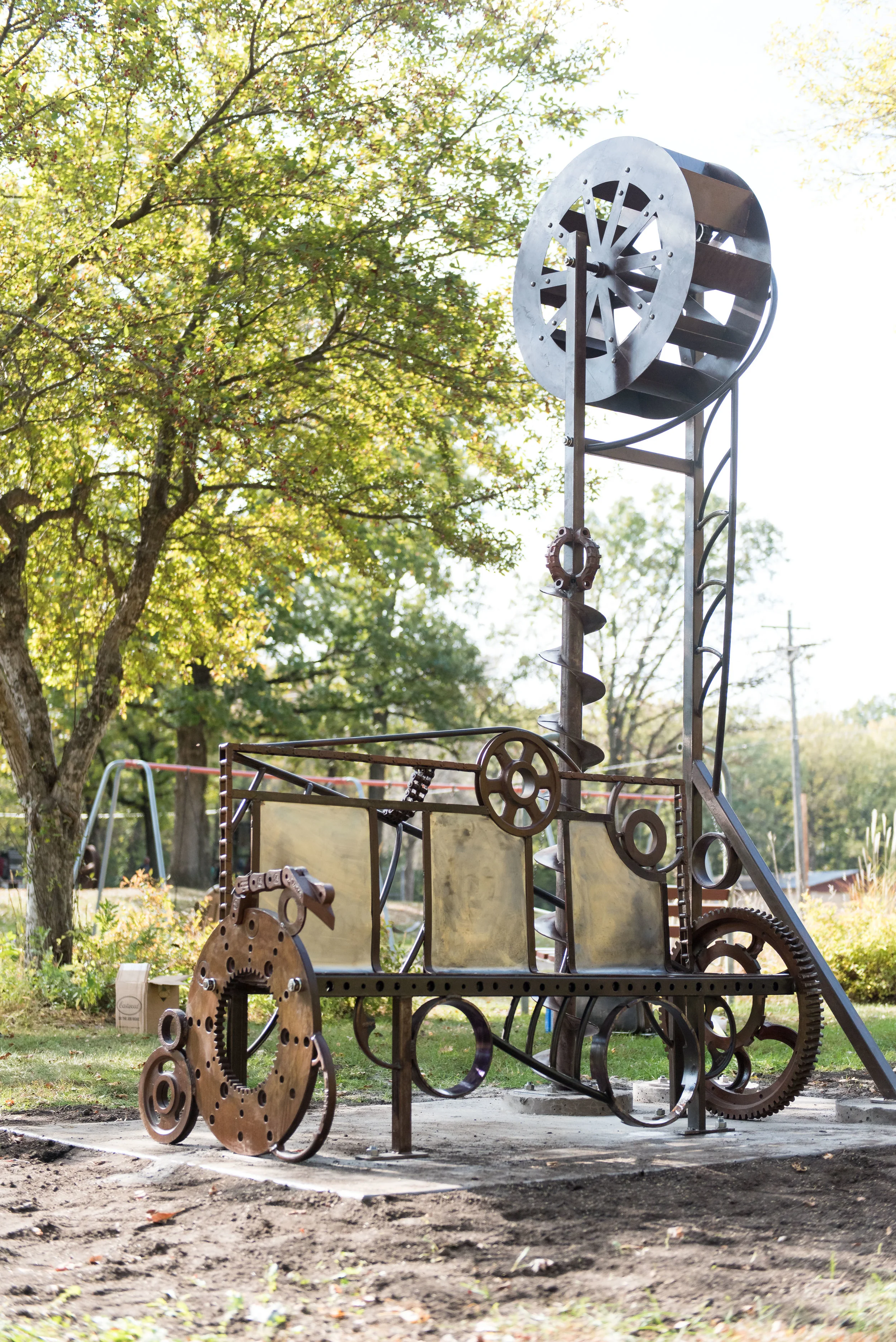 Metal sculpture resembling a motorcycle with gears, wheels, and mechanical parts, set outdoors with trees and a playground in the background.