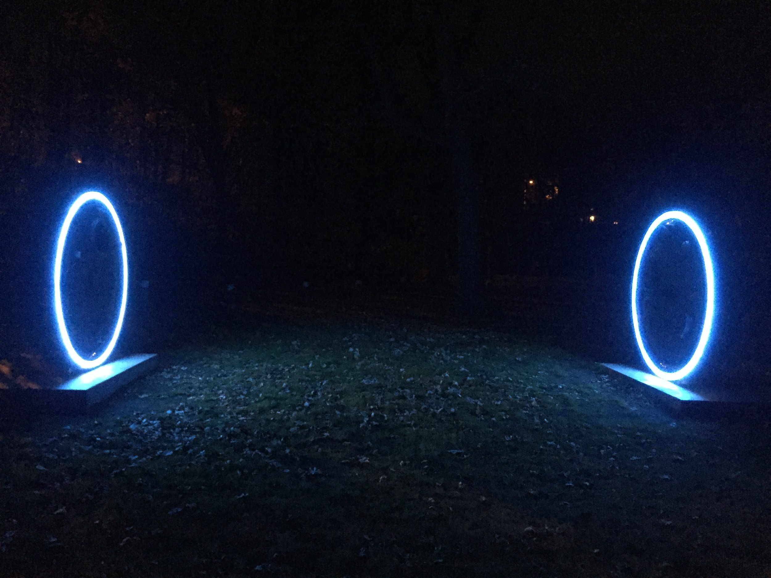 Nighttime outdoor scene featuring two illuminated circular sculptures with blue neon lights, positioned on rectangular bases, surrounded by dark trees and faintly lit background.