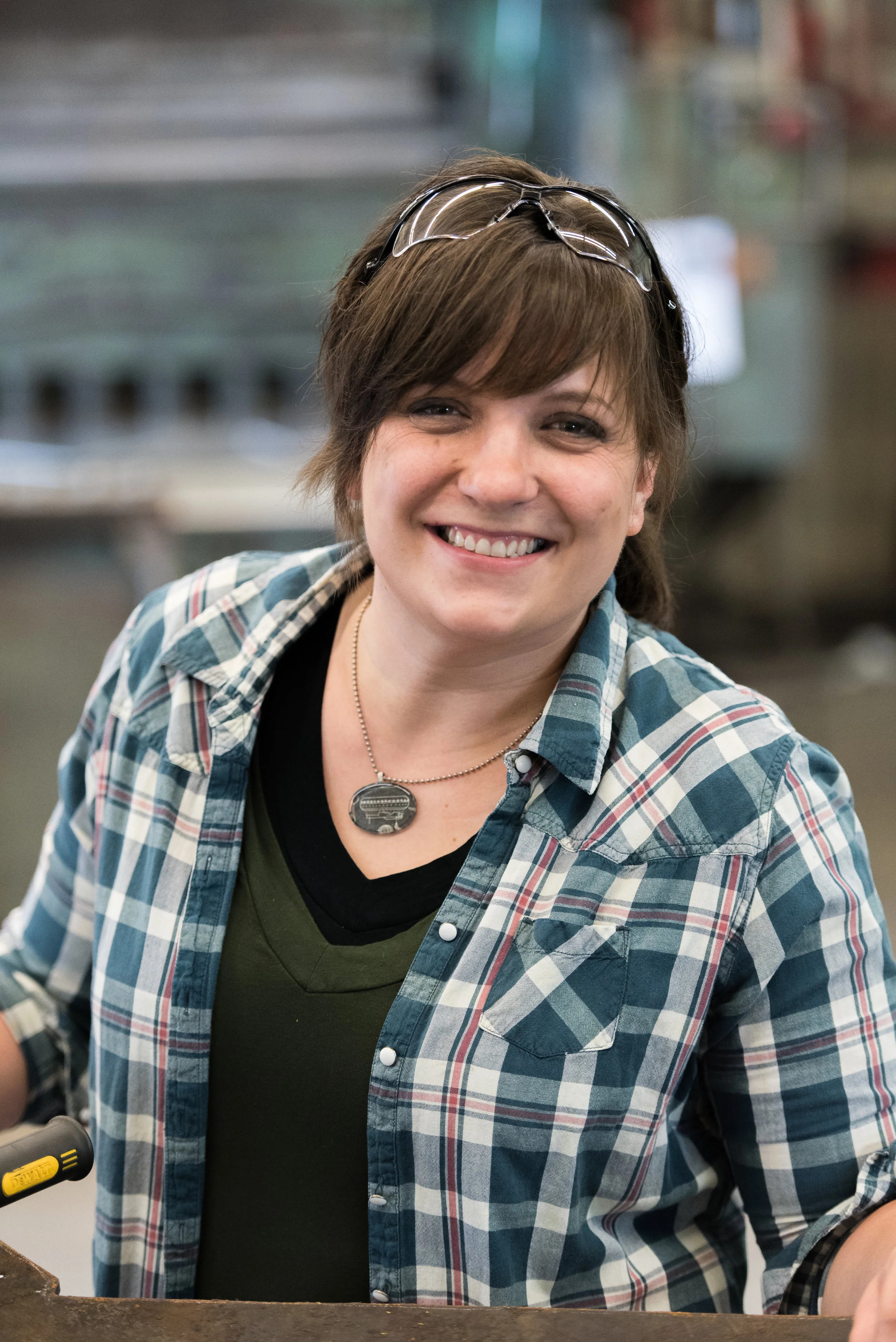 Woman with short brown hair wearing glasses on her head, smiling, wearing a green shirt and plaid button-up shirt, in a workshop or industrial setting.