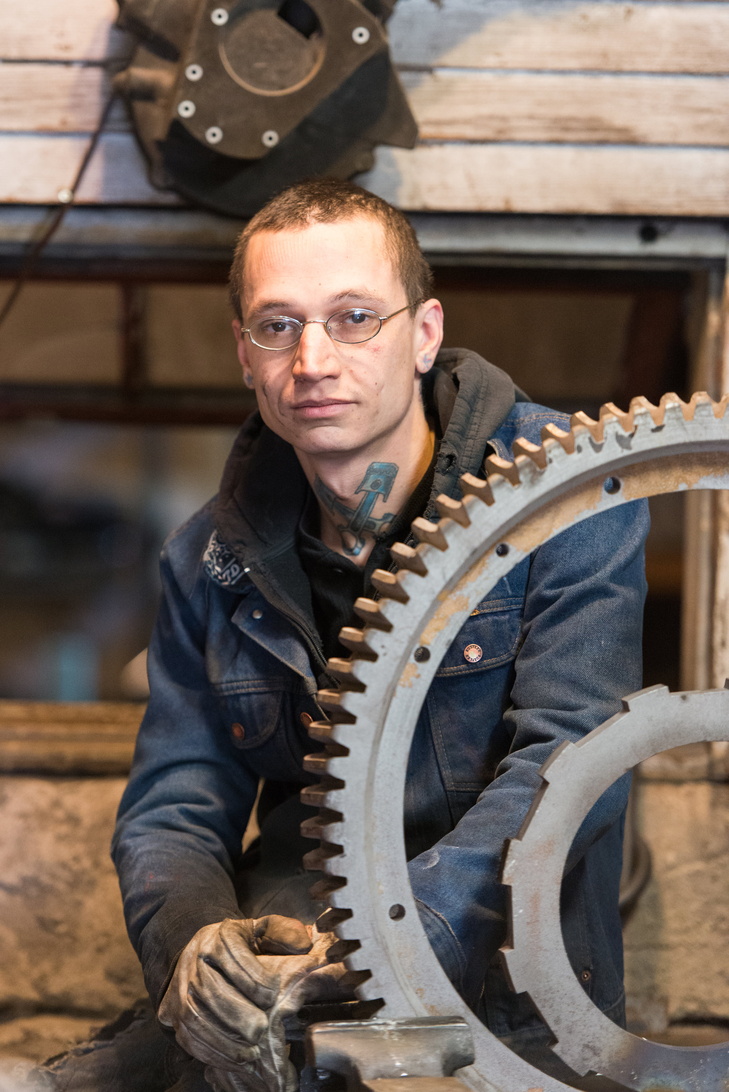 A young man with glasses, tattoos, and a denim jacket posing behind a large gear in a workshop or factory setting.