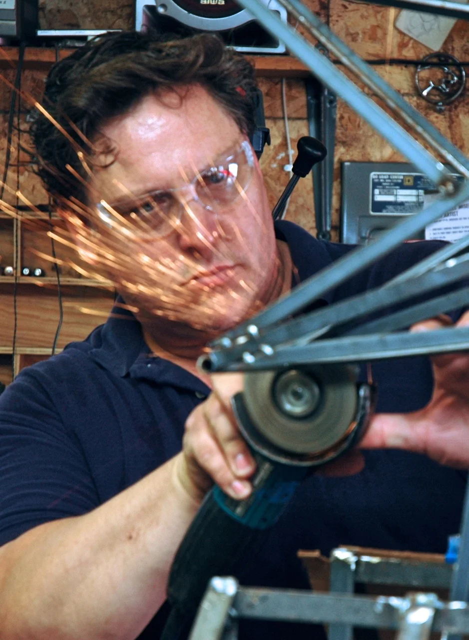 A man in safety glasses using a power tool, sparks flying, in a workshop setting with wooden walls and various equipment.