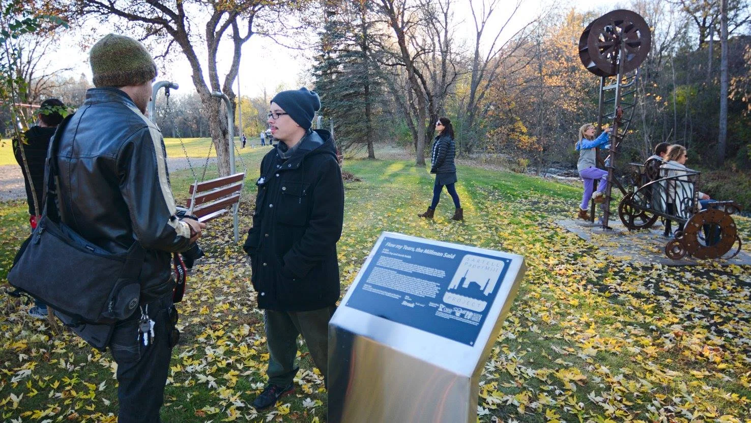 Two men talking in a park surrounded by trees and fallen leaves, with children playing on a metal sculpture and walking along a path.