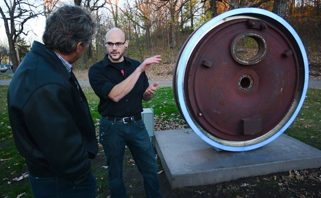 Two men standing outdoors near a large, rusted, circular object with a white neon border, possibly a billboard or art installation, during late afternoon or evening.