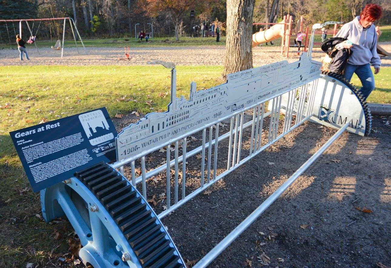 A small metal sculpture of a steamship with an informational plaque in front, located in a park with children playing on swings in the background.
