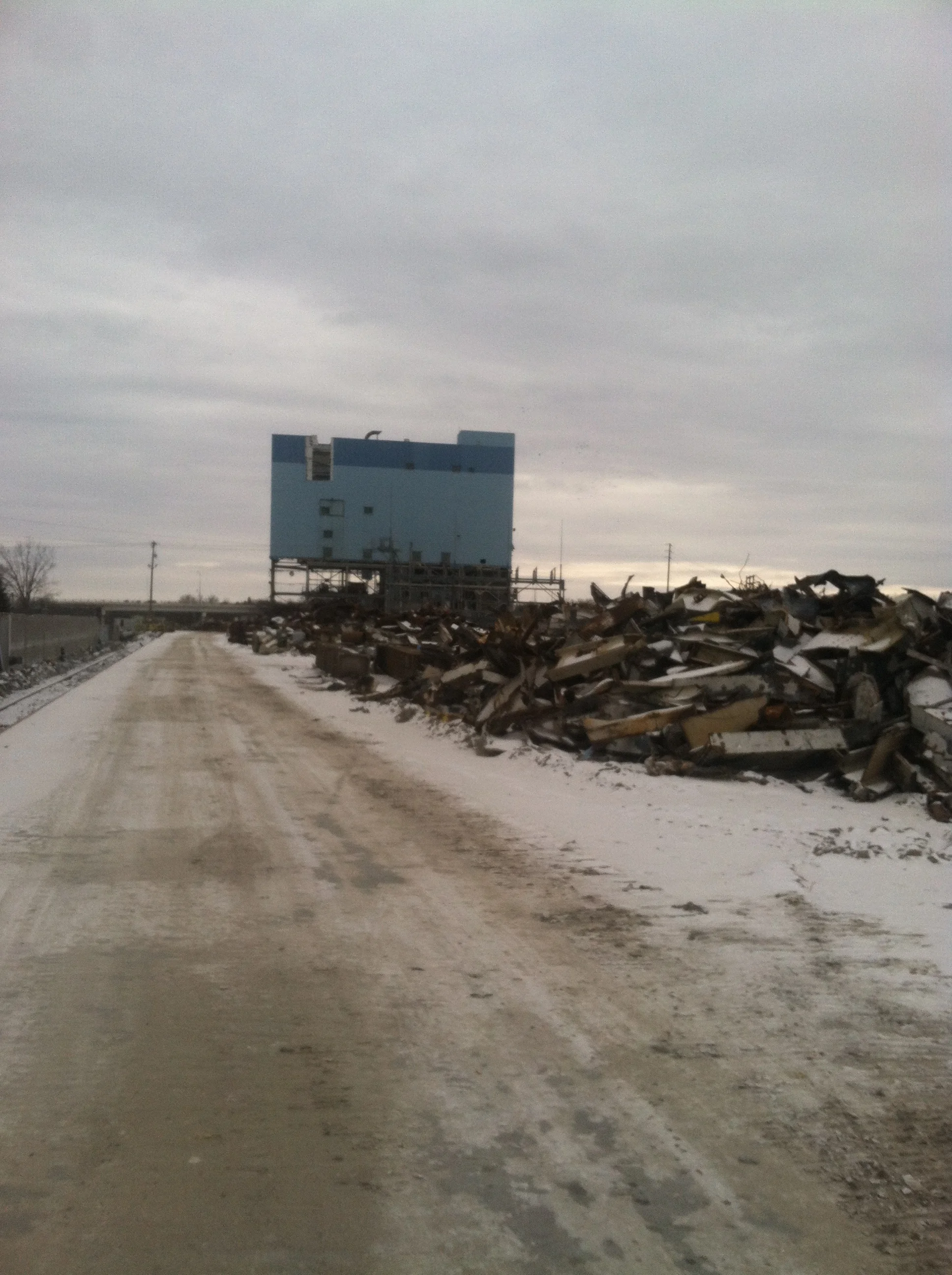 A dirt road runs alongside a pile of scrap metal and debris under a cloudy sky, with a blue building on stilts in the background.