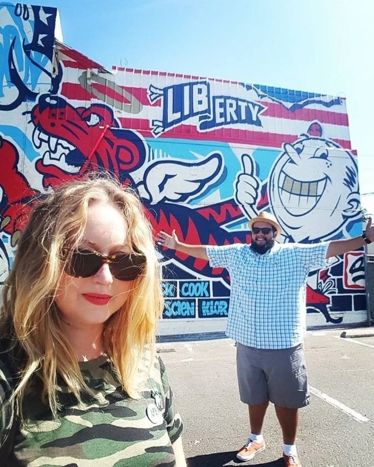 Two people taking a selfie in front of a colorful mural with the word "LIBERTY" and cartoon characters.