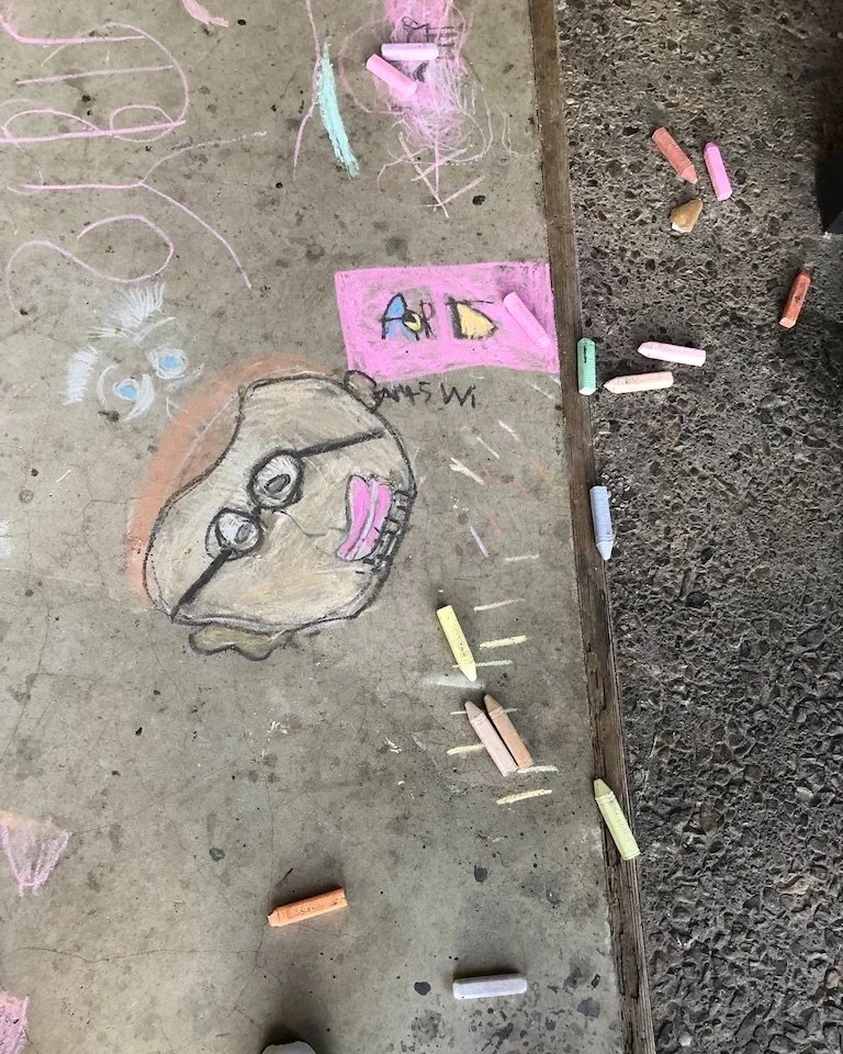 Colorful chalk drawings on concrete, including a large face with glasses, pink lips, and a small girl’s face, along with several small drawings and scribbles.