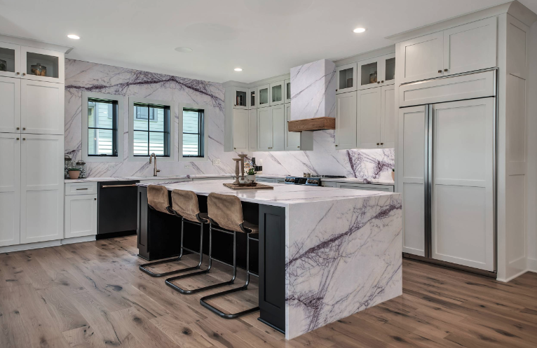 Cuisine moderne avec îlot central en marbre blanc avec veines violettes et meubles blancs, trois chaises en toile beige, plan de travail en marbre, fenêtres, sol en bois clair, et appareils intégrés.
