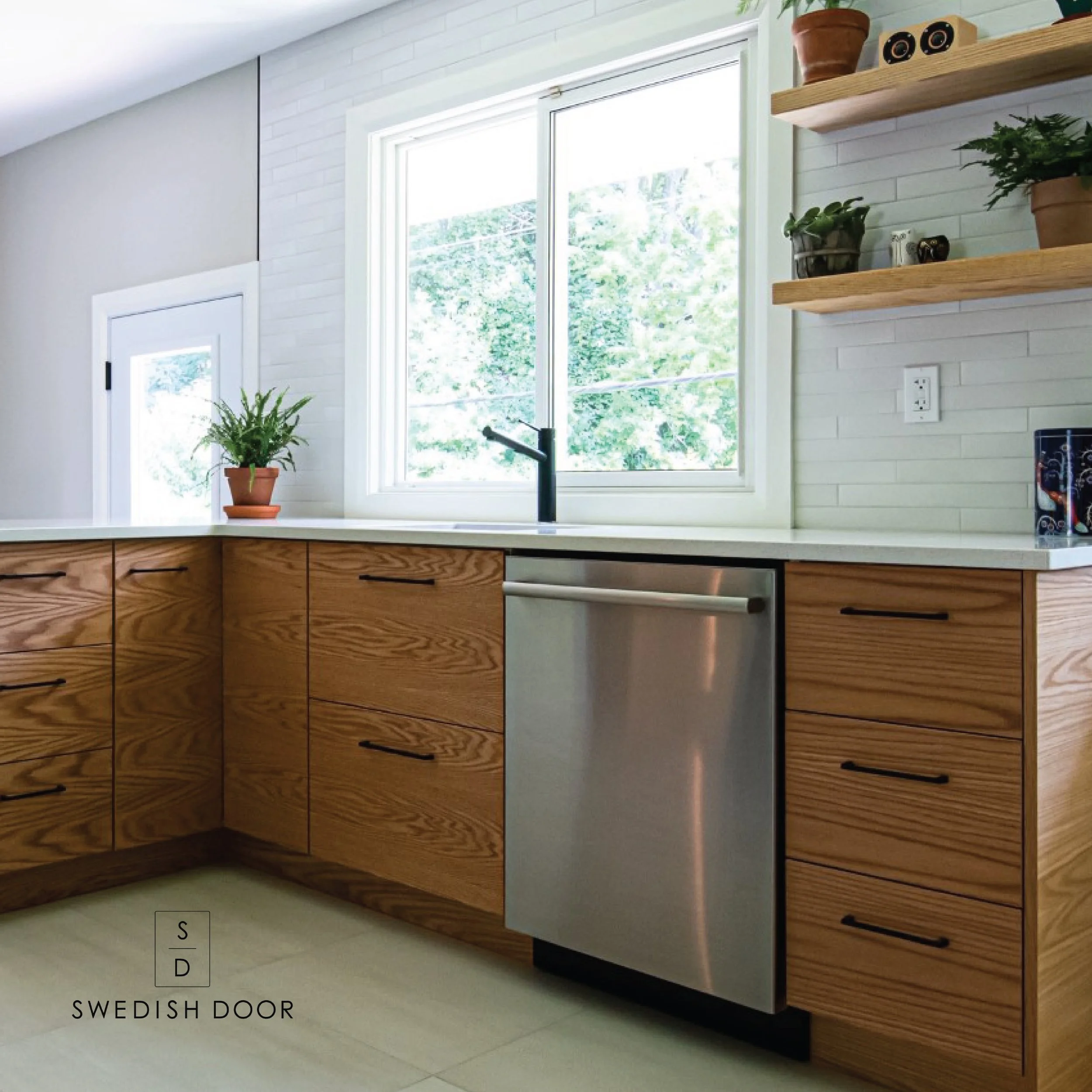 Cuisine avec fenêtres, mobilier en bois clair, un évier, une plante en pot sur le comptoir, étagères en bois avec plantes et objets décoratifs, prises électriques sur le mur.