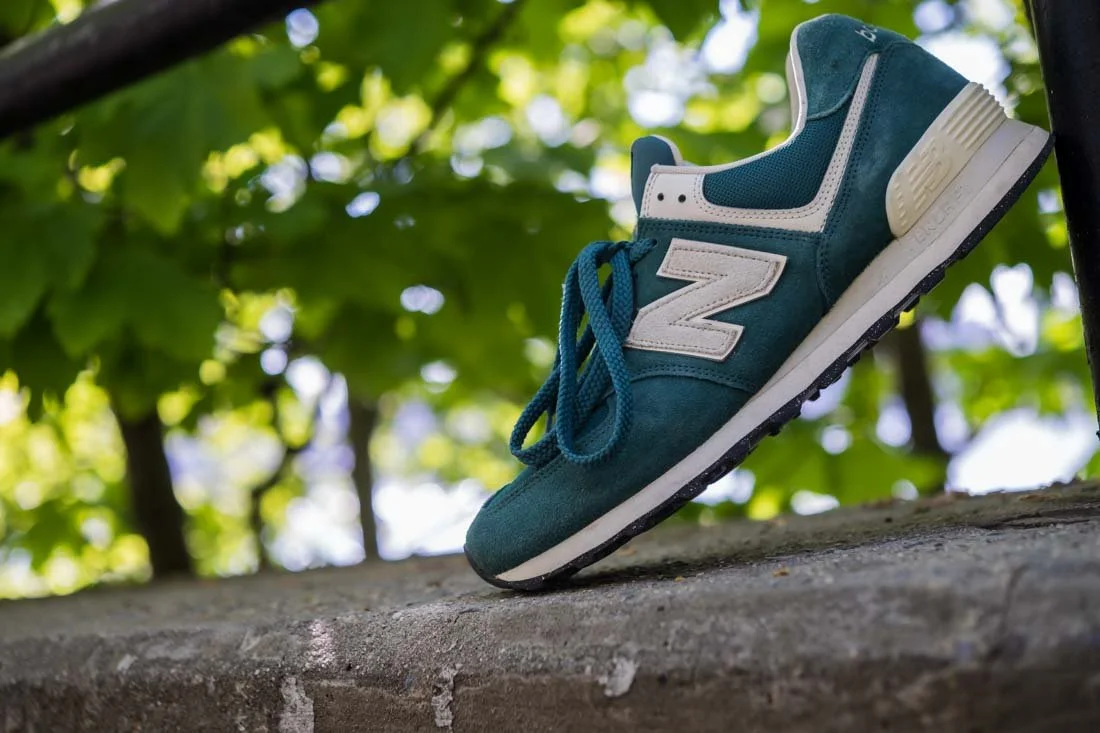 NEW BALANCE 574 COMFORT FOOTWEAR PHOTOS WIDE SHOT PROFILE LEANING ON FENCE