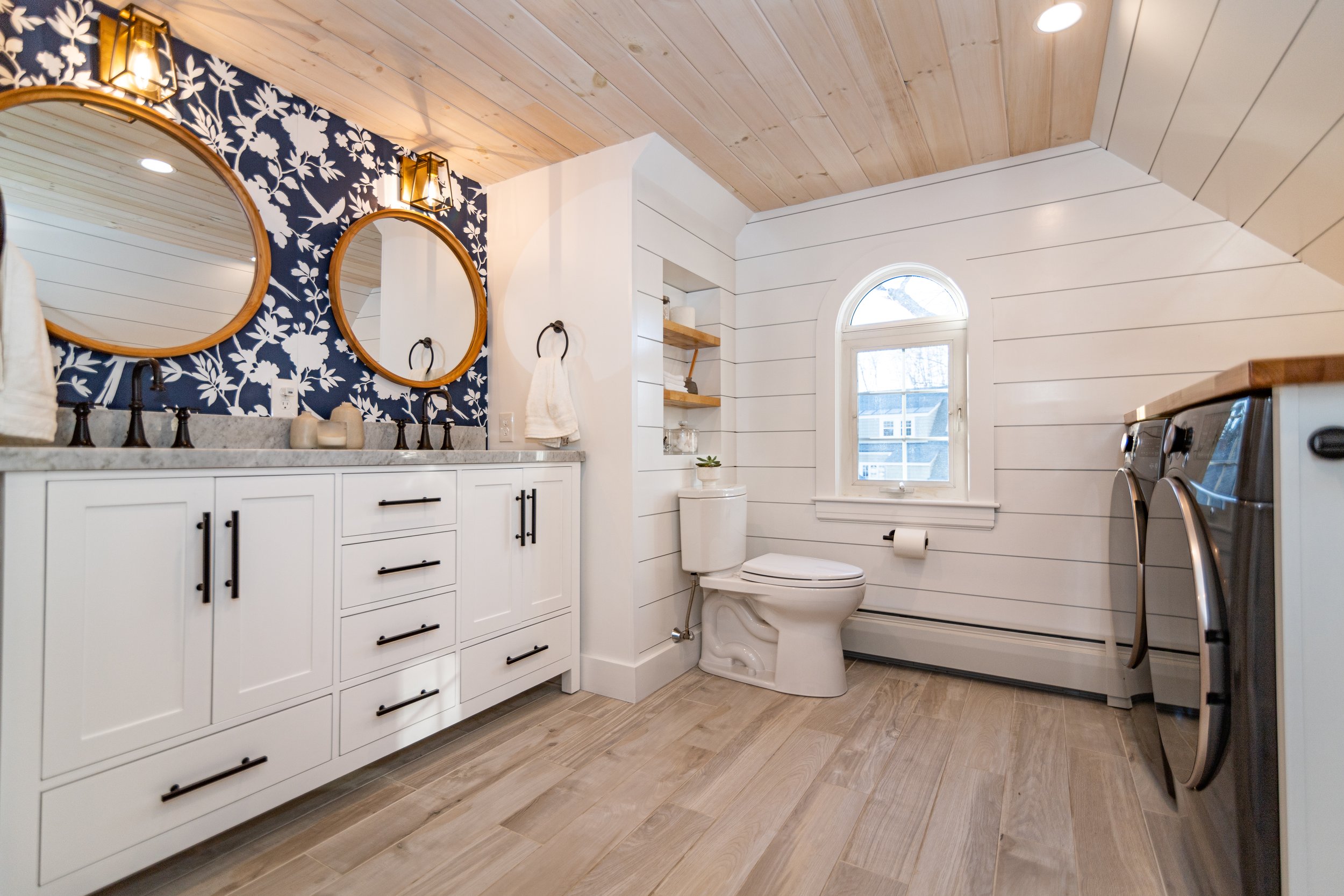 Bright attic bathroom featuring a white double vanity, navy floral wallpaper accent, wood flooring, built-in shelving, and a laundry area with black front-loading machines