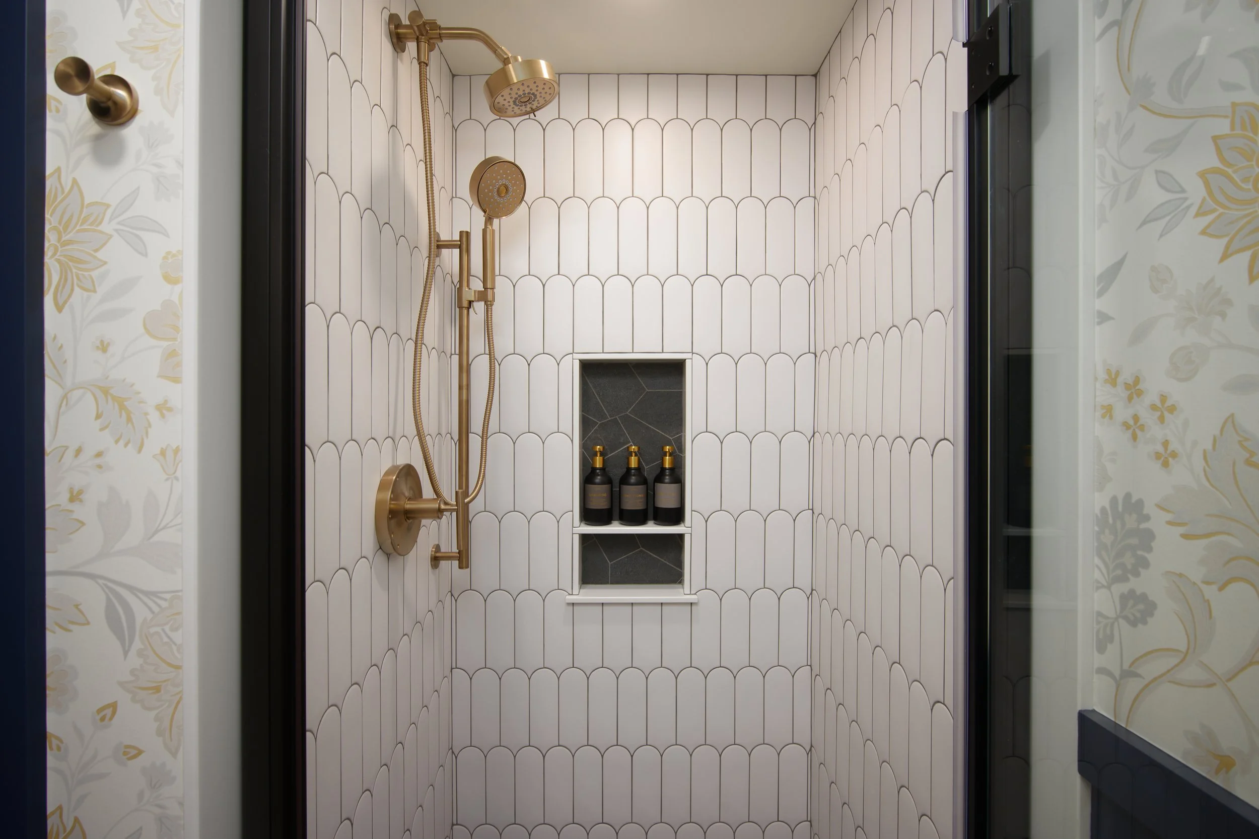 Walk-in shower with custom scalloped white tile, contrasting dark stone niches, and warm brass shower fixtures