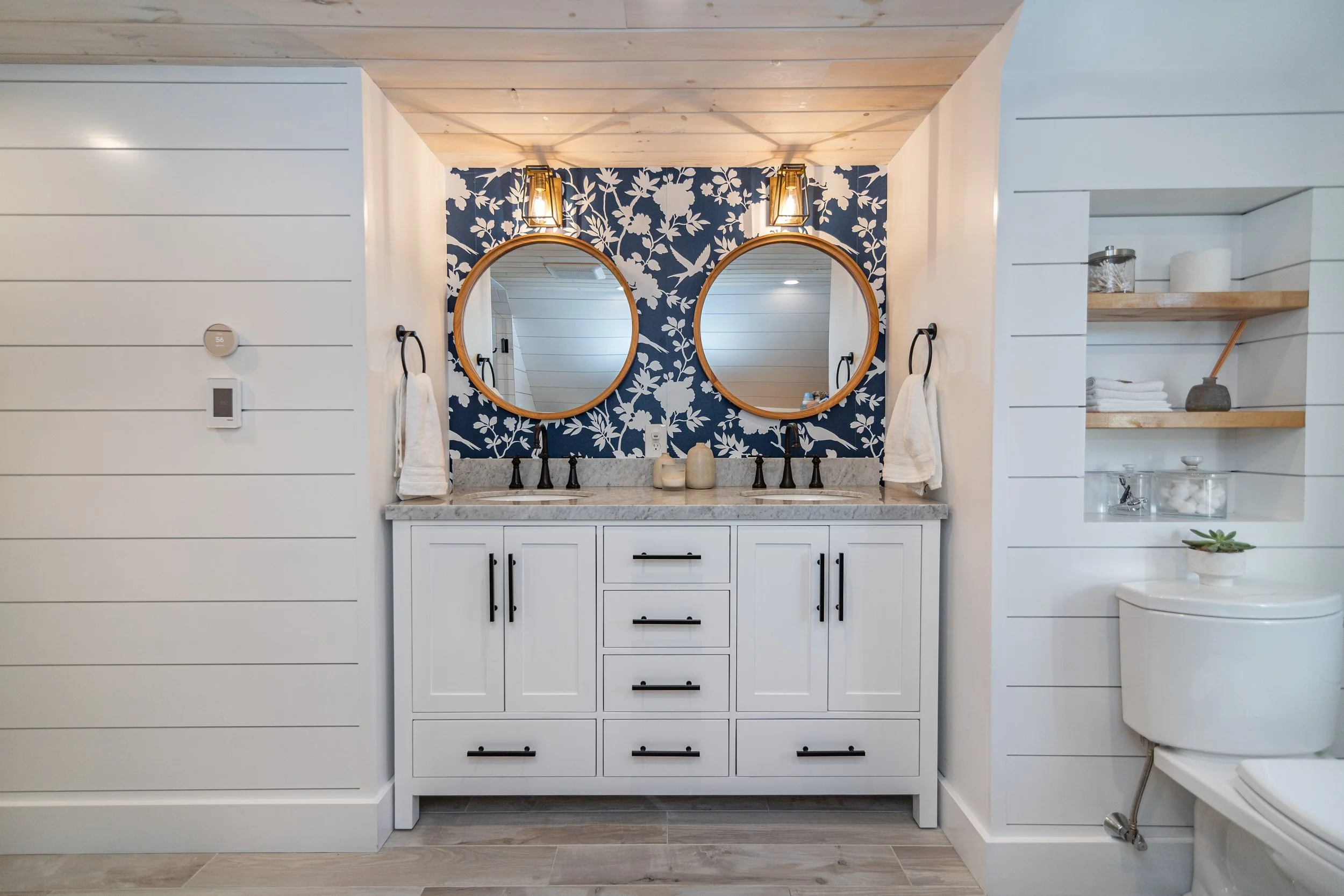 Stylish bathroom featuring a white double vanity, round wood-framed mirrors, floral wallpaper accent, and built-in shelving