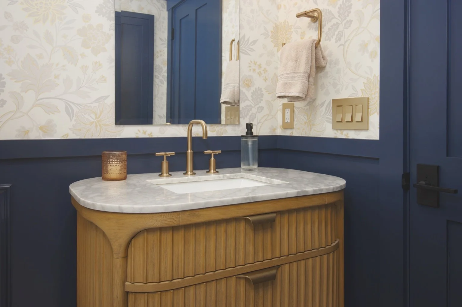 Natural oak vanity with a marble countertop, brass fixtures, navy wainscoting, and floral wallpaper in a remodeled powder room