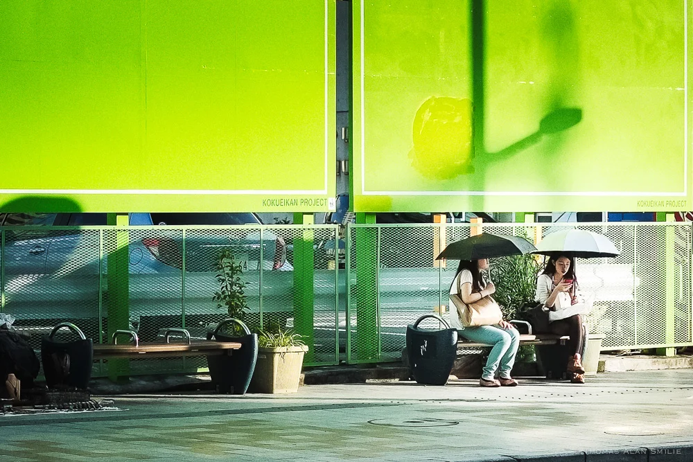Two women use their umbrellas to shield themselves from the sun while waiting for the bus on Kokusai Street in Naha. Fuji X-Pro1, 35mm f/1.4