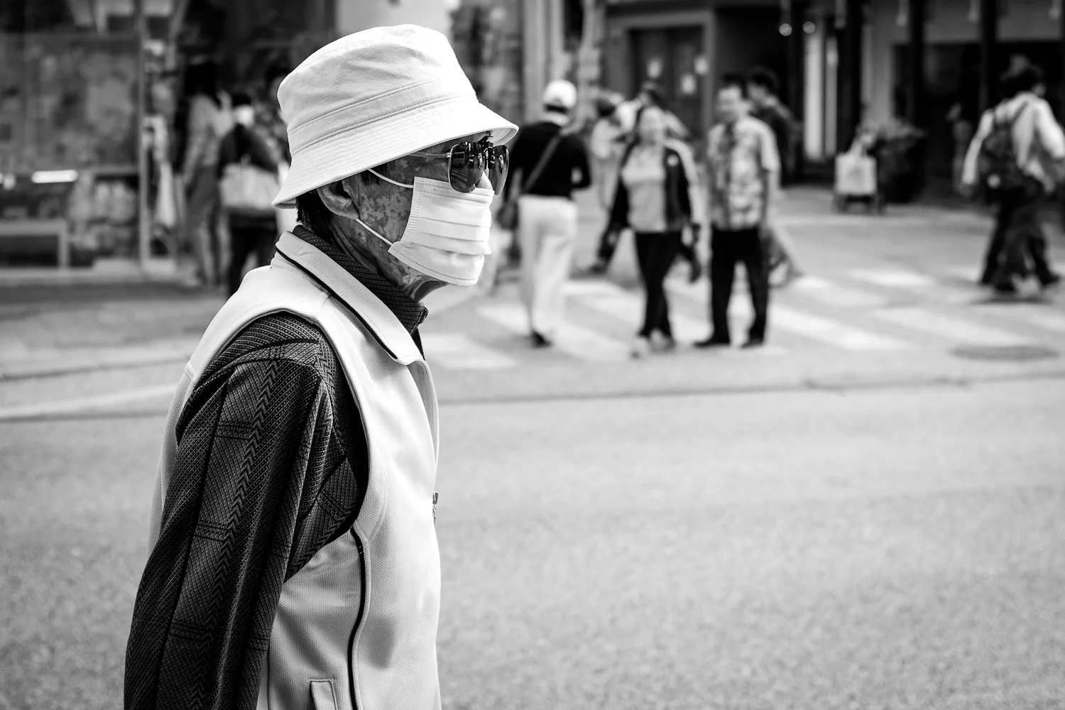 A man wearing a face mask, sunglasses and a hat walking on Kokusai street in Naha, Okinawa, Japan. Fuji x100s w/ TCLx100 Teleconverter at ISO200, f2.8, 1/500 sec.