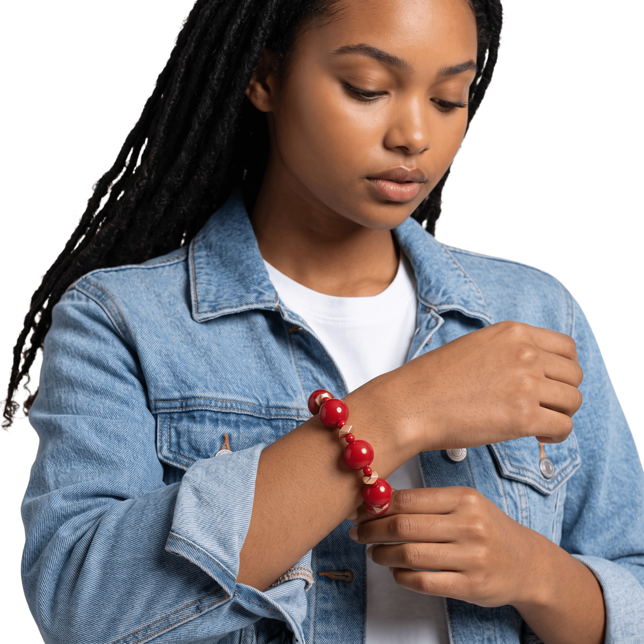 A young woman with braided hair and light brown skin, wearing a denim jacket and a red beaded bracelet, is holding her arm with her other hand.