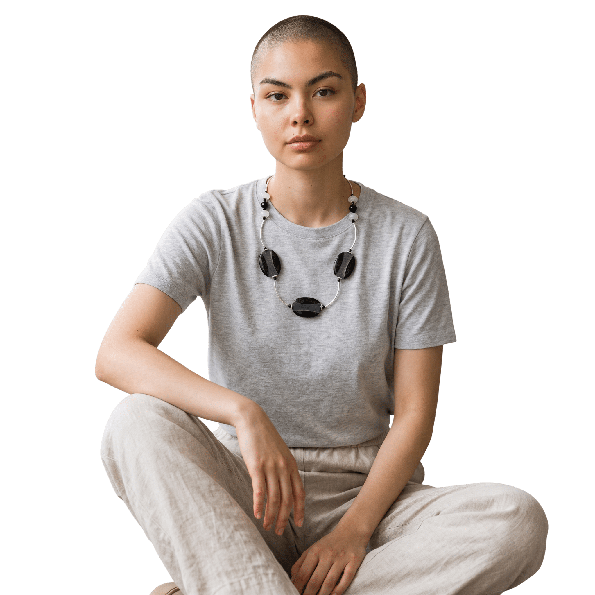 A young woman with a buzz cut hairstyle, wearing a gray t-shirt and beige pants, sitting with one knee up and wearing black wireless headphones around her neck, against a black background.