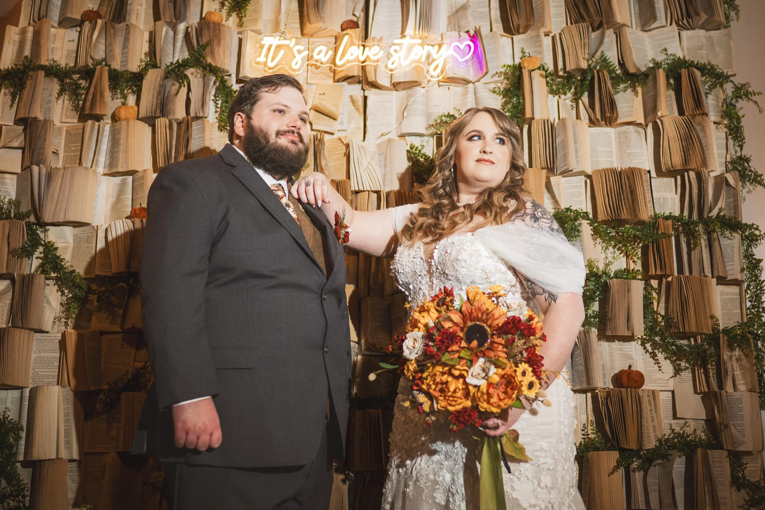 A bride and groom pose in wedding attire before a backdrop of open books and greenery, with a neon sign reading "It's a love story"—captured by a Northwest Indiana and Chicago wedding photographer.