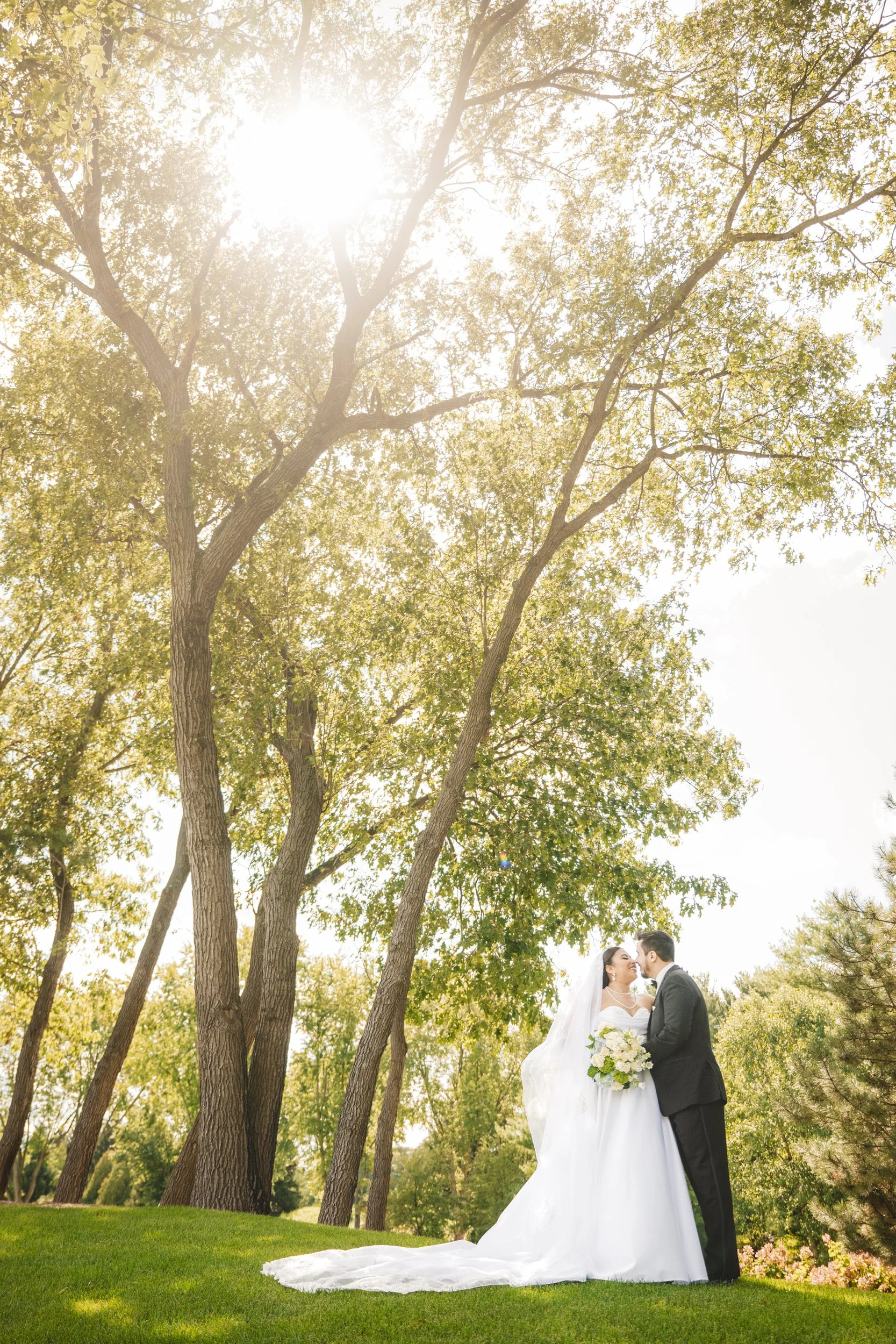 A bride and groom stand on green grass beneath tall trees, kissing under bright sunlight in an outdoor setting—captured beautifully by a northwest Indiana and Chicago wedding photographer.