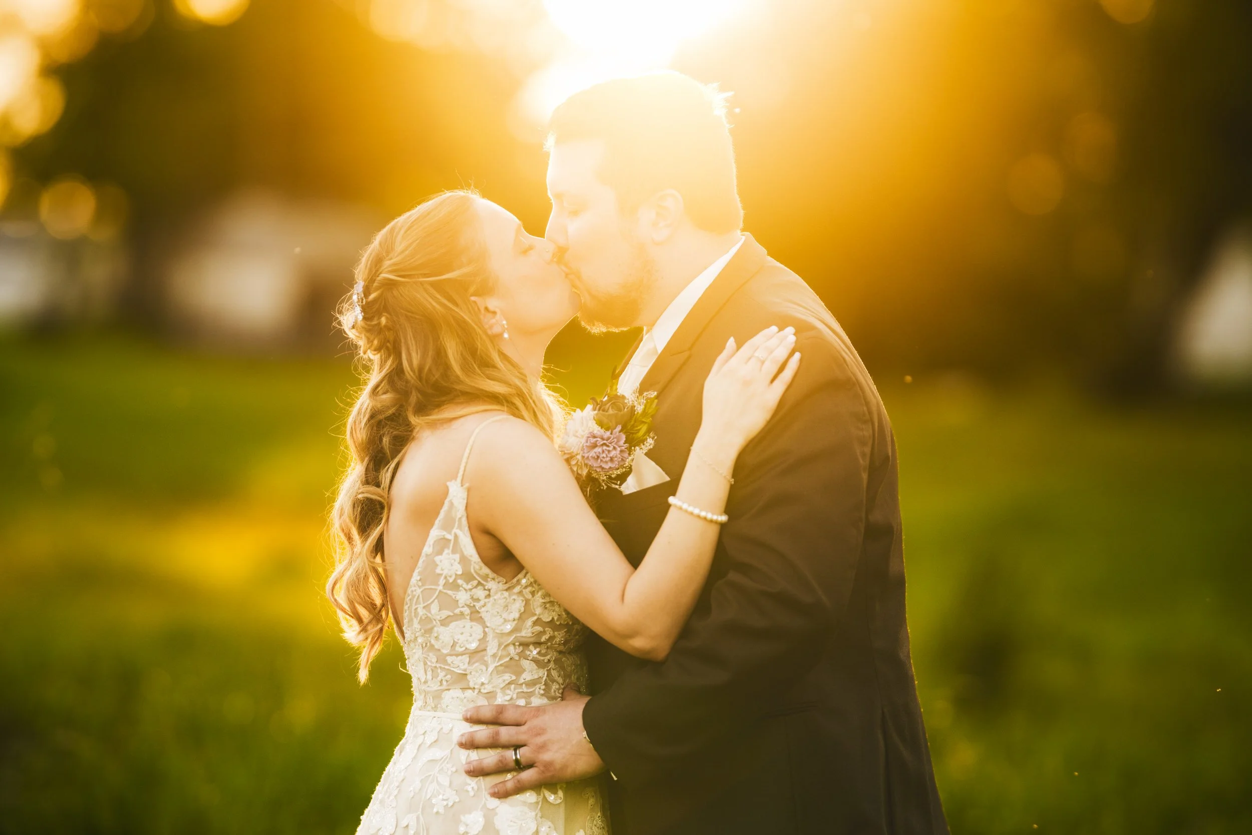 Bright golden hour sun streams upon a bride and groom kissing in a field behind The Design Barn in Hebron, Indiana.