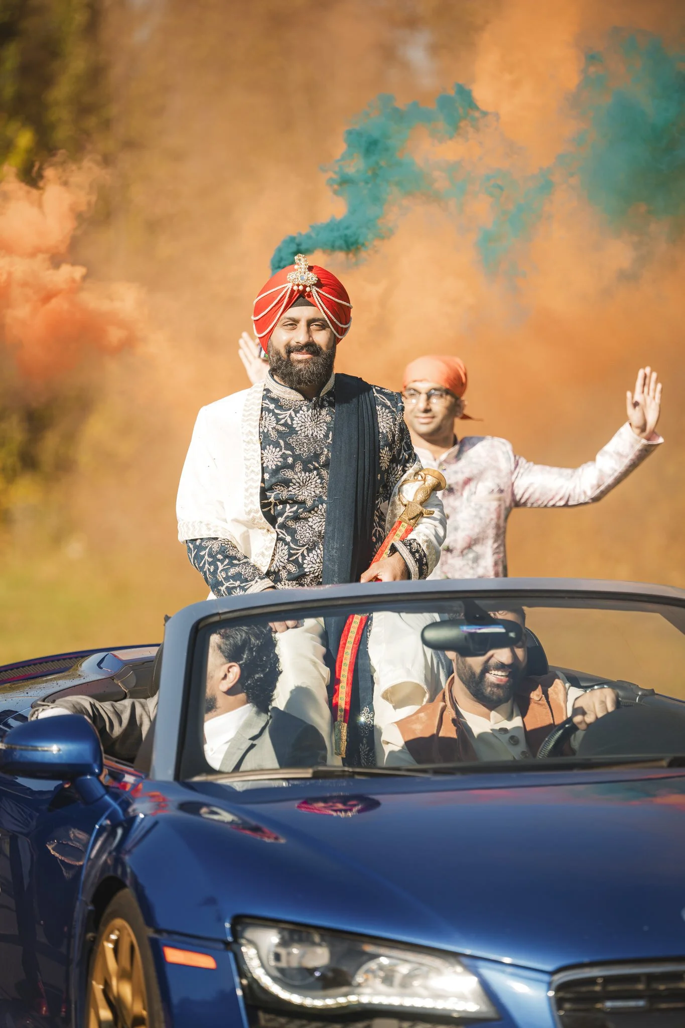 An Indian groom arrives in a convertible car with colored smoke bombs in the background.